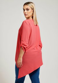 Blusa corallo con maniche 3/4, orlo curvo e tessuto testurizzato. Abbinata a jeans blu. Presenta un dettaglio con bottoni sulle maniche.