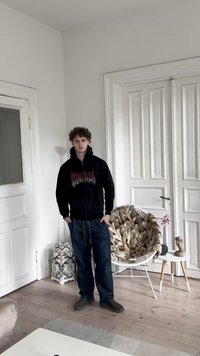 Sudadera negra con cremallera y gráfico multicolor, pantalones de sudadera azul oscuro con cordones, calzado negro, de pie en una habitación iluminada con paredes blancas.