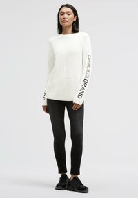 Weißer, gerippter Pullover mit langen Ärmeln und dem schwarzen Schriftzug "SOCXCXBRAND", kombiniert mit schwarzen Skinny-Jeans und schwarzen Sneakern.