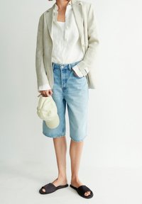 Vaaleanbeige pellavableiseri valkoisen paidan päällä, vaaleansiniset farkkushortsit, mustat liukusandaalit, kädessä vaaleankeltainen lippalakki. Rento kesäasu.