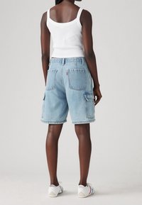 Mujer vestida con un shorts de cargo de mezclilla azul claro, una camiseta sin mangas blanca y zapatillas blancas, de pie contra un fondo gris liso, de espaldas a la cámara.