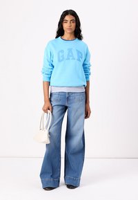 Sudadera azul claro con el logo "GAP", combinada con unos vaqueros de pierna ancha. Sosteniendo un pequeño bolso blanco, la modelo se encuentra de pie contra un fondo neutro.