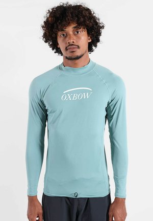 Homme aux cheveux bouclés portant un haut de sport bleu clair à manches longues avec le logo « OXBOW », debout devant un fond blanc uni.