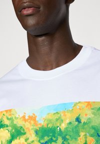 Hvid bomulds t-shirt med et farverigt, abstrakt landskabsdesign i grønne, orange og blå nuancer på forsiden.