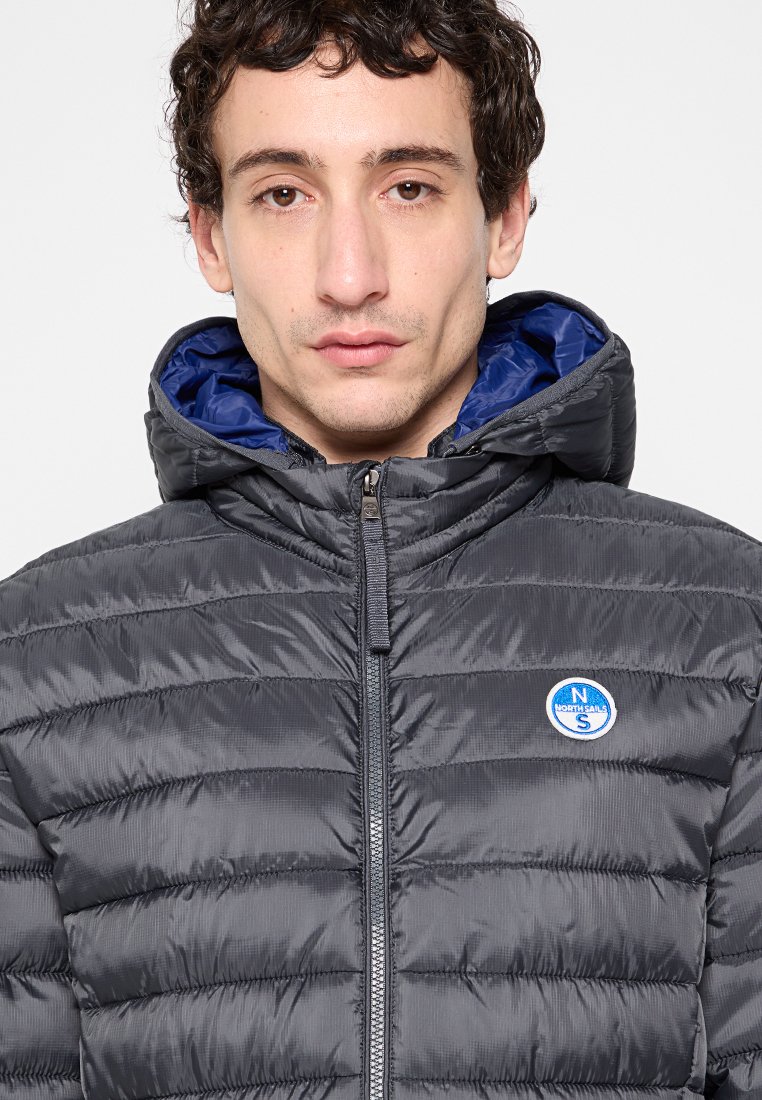 Giacca puffer grigia scura con foderatura interna blu, motivo trapuntato orizzontale, zip frontale e un logo circolare sul lato sinistro del petto.