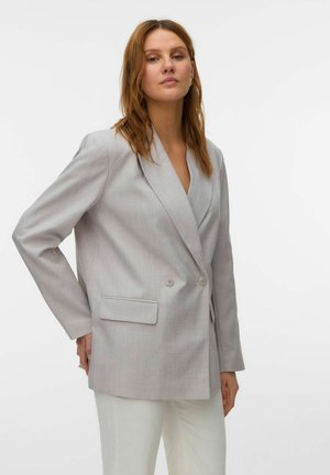 Femme aux cheveux châtain clair portant un blazer gris clair à double boutonnage et un pantalon blanc, debout devant un fond uni.