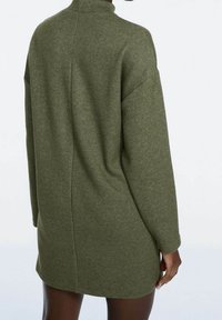 Henkilö pukeutuneena polvimittaisessa, pitkähihaisessa oliivinvihreässä neulemekossa, esitettynä selkäpuolelta yksinkertaista valkoista taustaa vasten.