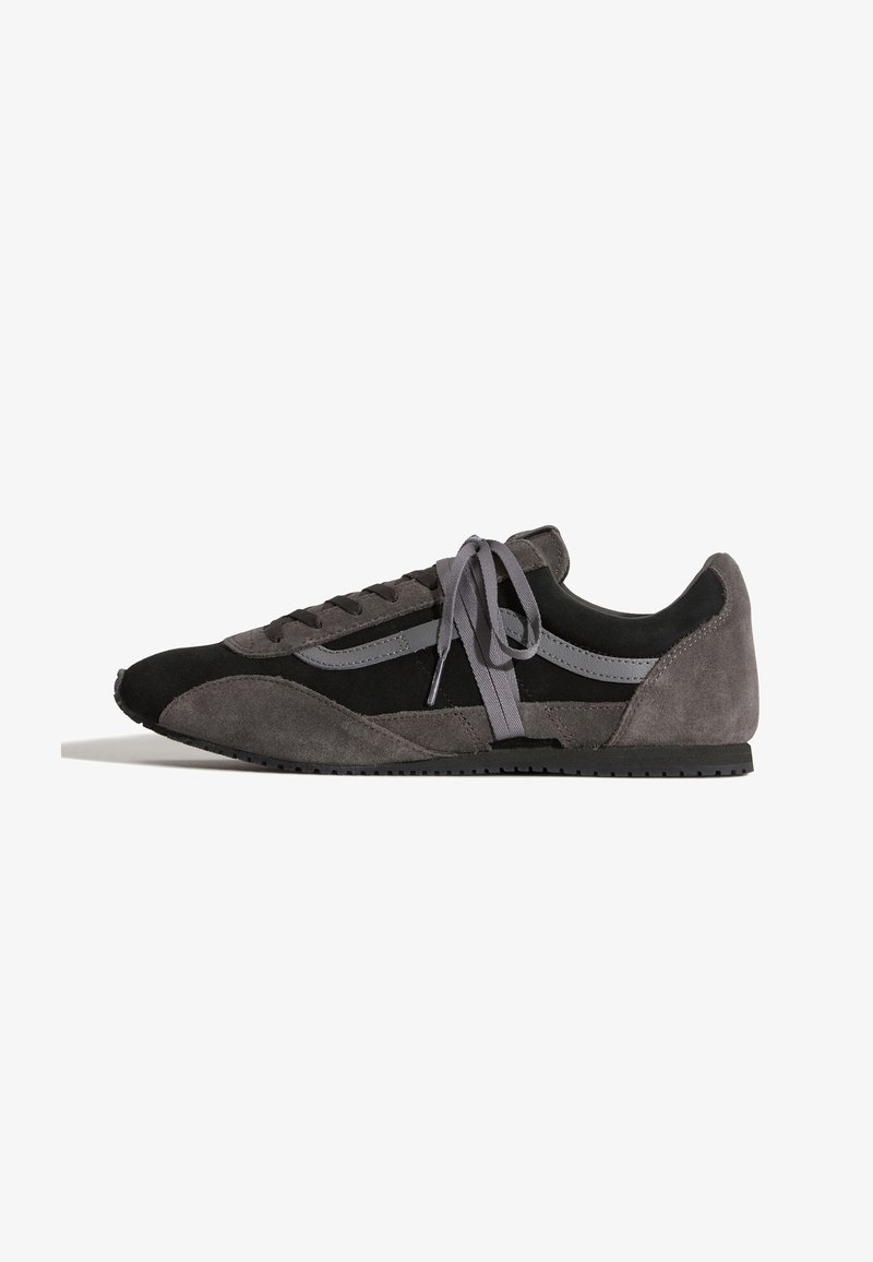 Μαύρο και σκούρο γκρι χαμηλό sneaker με σουέτ και υφασμάτινα πάνελ, γκρι κορδόνια δεμένα σε φιόγκο και επίπεδη μαύρη σόλα.