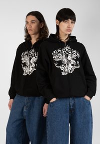 Black zip-up hoodies feature white graphic designs. Paired with loose blue denim pants, the overall look emphasizes bold contrasts and textures.