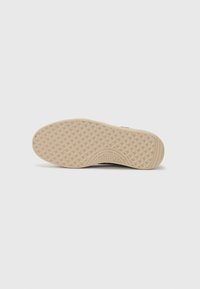 Suola della scarpa con un design in gomma beige testurizzata, caratterizzata da un motivo a diamante e un logo impresso. Forma piatta e arrotondata per una maggiore adattabilità.