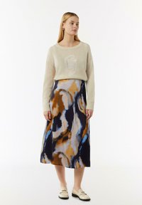 Pull beige avec un logo texturé, associé à une longue jupe fluide ornée d'un motif tourbillonnant dans des tons de bleu, marron et noir.