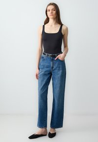 Body nero senza maniche abbinato a jeans blu larghi a vita alta. Cinturato in vita, indossa delle ballerine nere. Sfondo semplice.