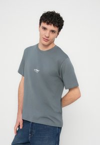 Szürke pamut póló rövid ujjakkal, kerek nyakkivágással és egy kis fehér logóval a mellkason. Kék farmernadrággal viselve.