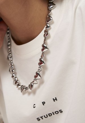 Close-up van een zilveren hartvormige kraal halsketting die over een witte top wordt gedragen met "CPH STUDIOS" in zwarte letters gedrukt.