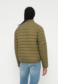 Chaqueta de plumas verde oliva con acolchado horizontal, cuello alto y detalles acanalados en los hombros. Llevada con unos vaqueros azul claro.