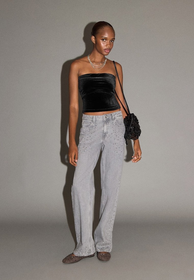 Top negru fără bretele din catifea, blugi largi din denim gri deschis cu accente cu ținte și o geantă texturată neagră. Fundal gri.