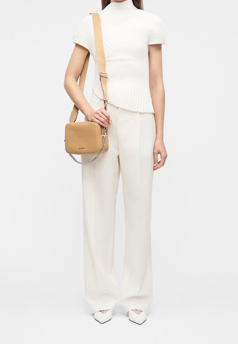 Femme portant un haut blanc côtelé à manches courtes et un pantalon large, tenant un sac bandoulière carré beige avec détail chaîne.