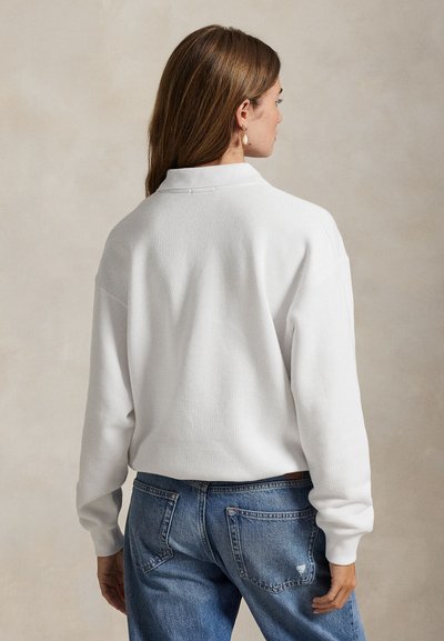 Pikade pruunide juustega naine, kes kannab valget pika varrukaga särki ja siniseid teksapükse, näoga neutraalse tausta poole.