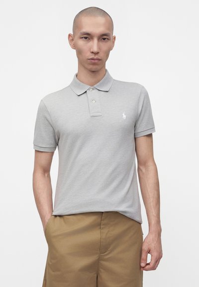 Jeune homme aux cheveux courts portant un polo gris clair et un short beige, debout, une main dans la poche, sur un fond uni.