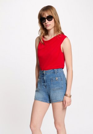 Donna che indossa occhiali da sole neri, top rosso senza maniche, shorts di jeans blu a vita alta con grandi tasche frontali, e braccialetti al polso destro.