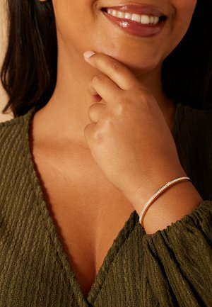 Gold bangle bracelet with a row of small clear stones, worn on a bare wrist, against a textured olive green fabric.