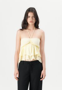 Top galben stratificat fără bretele, cu o margine volanată și detalii strânse în centrul, purtat cu pantaloni negri. Material neted și ușor.
