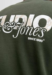 Tmavě zelené bavlněné tričko s bílým textem a texturou v maskáčovém vzoru "Audio & Jones" a "Od roku 1990" na zadní straně.