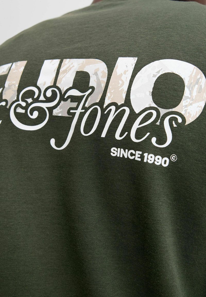 Tmavě zelené bavlněné tričko s bílým textem a texturou v maskáčovém vzoru "Audio & Jones" a "Od roku 1990" na zadní straně.