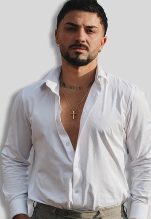 Weißes Hemd mit offenem Kragen, hellbeige Hose, goldene Kreuzkette und sichtbare Tattoos, schwarze Haare und Bart.
