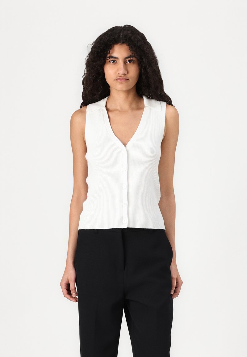 Haut sans manches blanc à côtes avec un décolleté en V profond et fermeture à boutons. Texture lisse et design ajusté, porté avec un pantalon noir.