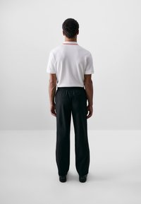 Chemise polo blanche à manches courtes avec des accents de col rouges et un pantalon noir à taille élastique, vue de dos contre un fond uni.