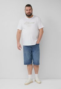 Biała bawełniana koszulka z złotym nadrukiem, zestawiona z niebieskimi denimowymi szortami. Model nosi białe sneakersy i skarpetki do kostek. Prosty, codzienny strój.