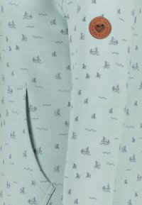 Sudadera verde claro de tela suave, con un patrón repetido de velas y un parche de logo marrón en la manga. Bolsillo visible en la parte frontal.