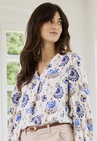 Femme aux longs cheveux bruns portant un chemisier blanc à motifs floraux bleus et un pantalon beige, debout à l'intérieur près d'une fenêtre.