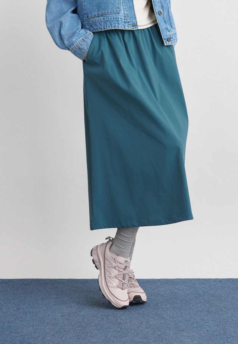 Spodní polovina osoby oblečené v tyrkysové midi sukni, šedých ponožkách, růžových teniskách a džínové bundě s rukama v kapsách.