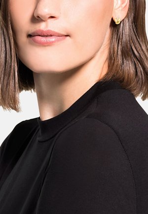 Mujer con cabello corto castaño que lleva pequeños pendientes de nudo dorados y una blusa negra de cuello alto, mostrada desde la barbilla hasta el hombro.