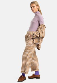 Maglioncino violaceo con colletto alto, pantaloni a quadri marrone a vita alta, scarpe marroni e calze viola, con il maglioncino leggermente appoggiato sul braccio.