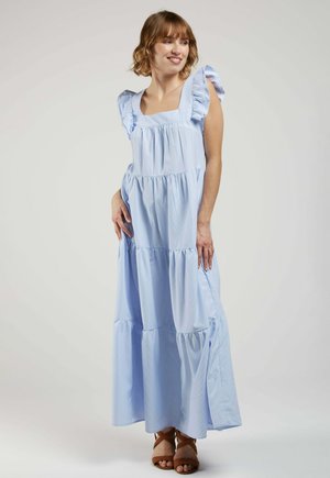 Femme souriante portant une robe maxi sans manches bleu clair à étages avec des bretelles à volants et des sandales marron à lanières, debout sur un fond uni.