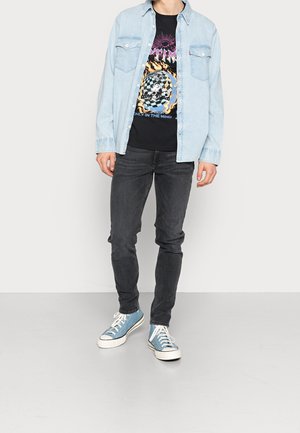 Camisa de mezclilla azul clara superpuesta a una camiseta gráfica negra, combinada con jeans oscuros de corte ajustado y zapatillas de lona azules con suelas blancas.