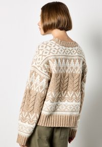 Gruby sweter z dzianiny z kremowymi i beżowymi wzorami, w tym warkoczami i geometrycznymi kształtami. Ściągaczowy dół oraz szczegółowe, teksturowane rękawy.