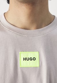 T-shirt en tissu gris avec un patch rectangulaire vert fluo portant le texte "HUGO" en lettres noires en gras, centré.