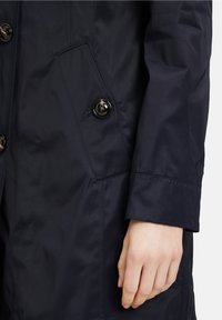 Manteau imperméable bleu marine avec une texture lisse, une fermeture à double bouton, des poches latérales et des poignets cousus, mettant en avant des détails de conception fonctionnels.