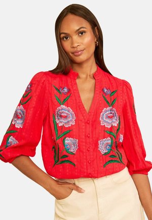 Kvinde iført en rød bluse med broderede blå og pink blomster samt grønne stilke, kombineret med lyse beige bukser, poserende med den ene hånd i lommen.
