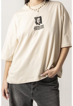 Kobieta w beżowej, oversize'owej koszulce z czarną grafiką twarzy i napisem, w połączeniu z perłowym naszyjnikiem i ciemnoszarymi dżinsami, z ręką w kieszeni.