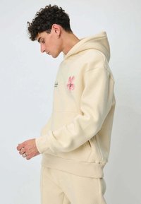 Hoodie beige avec une poche avant, arborant un motif de fleur rose clair et un petit texte noir. Tissu doux avec poignets et ourlet côtelés.