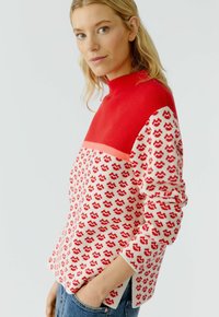 Rot und cremefarben gemusterter Pullover mit hohem Kragen, langen Ärmeln und aufgespaltenem Saum. Das Design enthält kleine, rotwellenartige Motive.