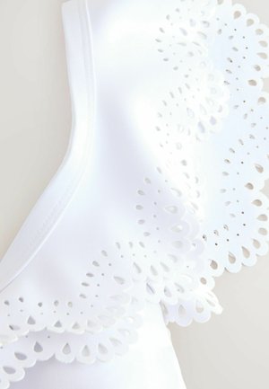 Bílý oděv s propracovanými výřezy podél okrajů, který má hladkou texturu a strukturovaný design pro přiléhavý vzhled.