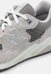 Szürke sneaker, textúrált velúr felsőrésszel, hálós részletekkel, kontrasztos logóval és fehér fűzőkkel. Gumi talp, barázdált mintázattal.