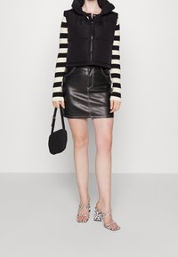 Czarna kamizelka puchowa z kapturem, paskowana bluzka z długim rękawem, czarna skórzana mini spódnica i sandały na wysokim obcasie w szachownicę, trzymająca małą czarną torebkę.