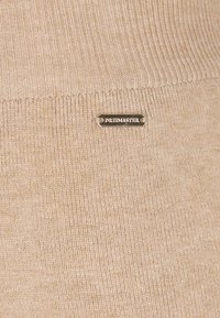 Beige neulottu neulepusero, jossa on ribbikuvio ja pieni metallilogo, jossa lukee "DREIMASTER", edessä.